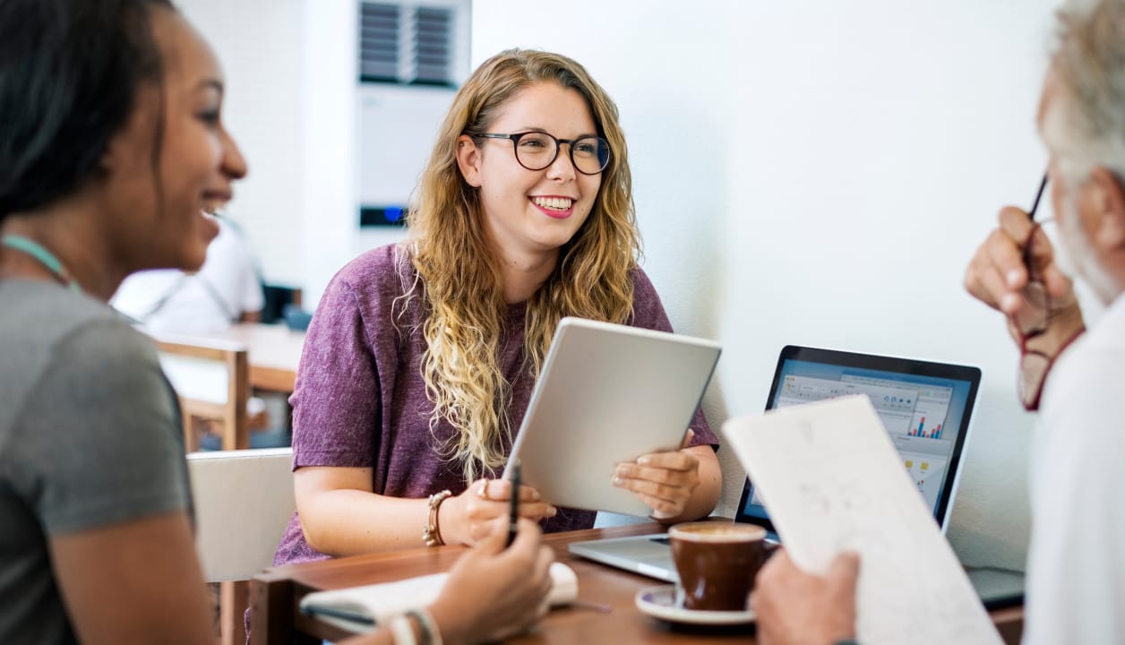 A group of students smiling and working together at a table with a tablet and laptop.