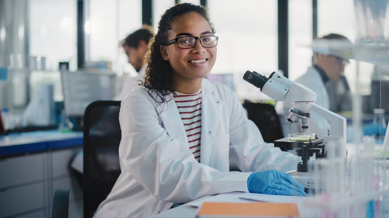 female scientist sits in a lab in front of a microscope smiling