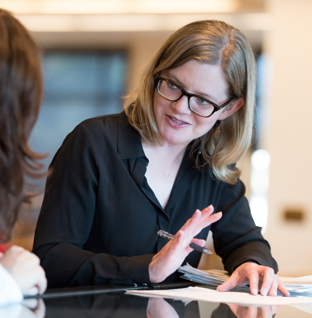 A woman with glasses and blonde hair speaking and gesturing during a meeting.