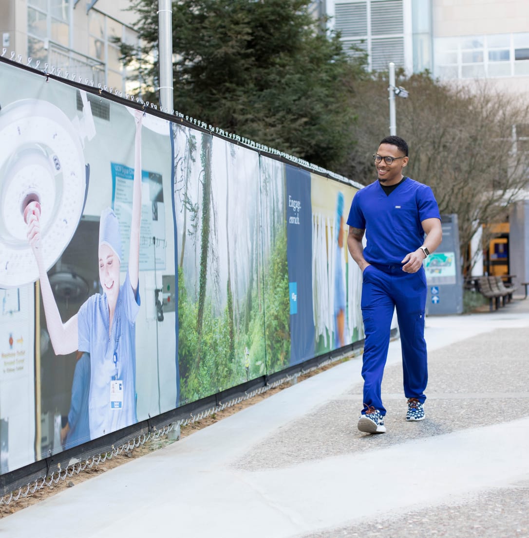 A smiling man in blue medical scrubs walking along an outdoor campus path.