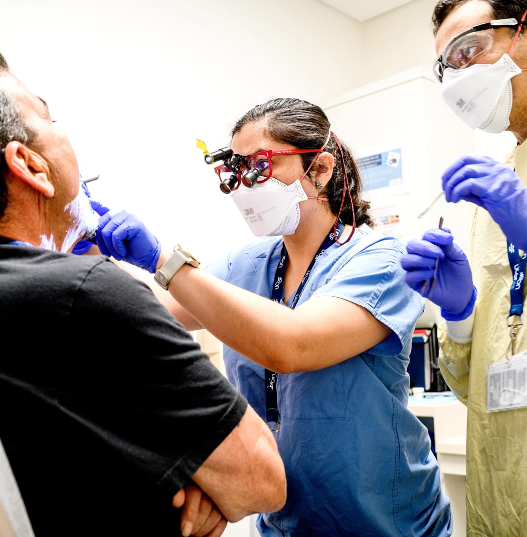 A dental professional wearing magnifying loupes and a face mask performs a procedure on a patient.