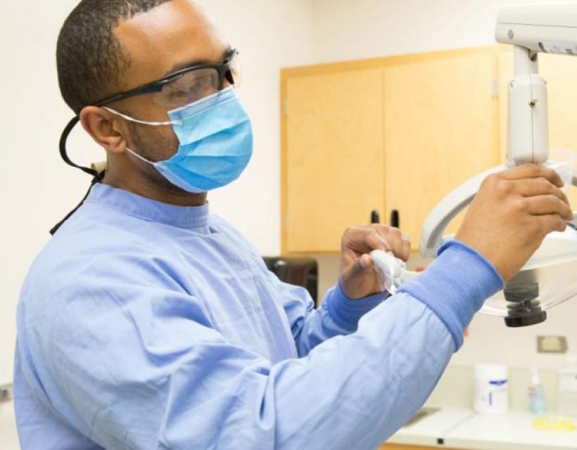 A healthcare professional wearing a blue gown, face mask, and safety glasses adjusting an overhead clinic light.